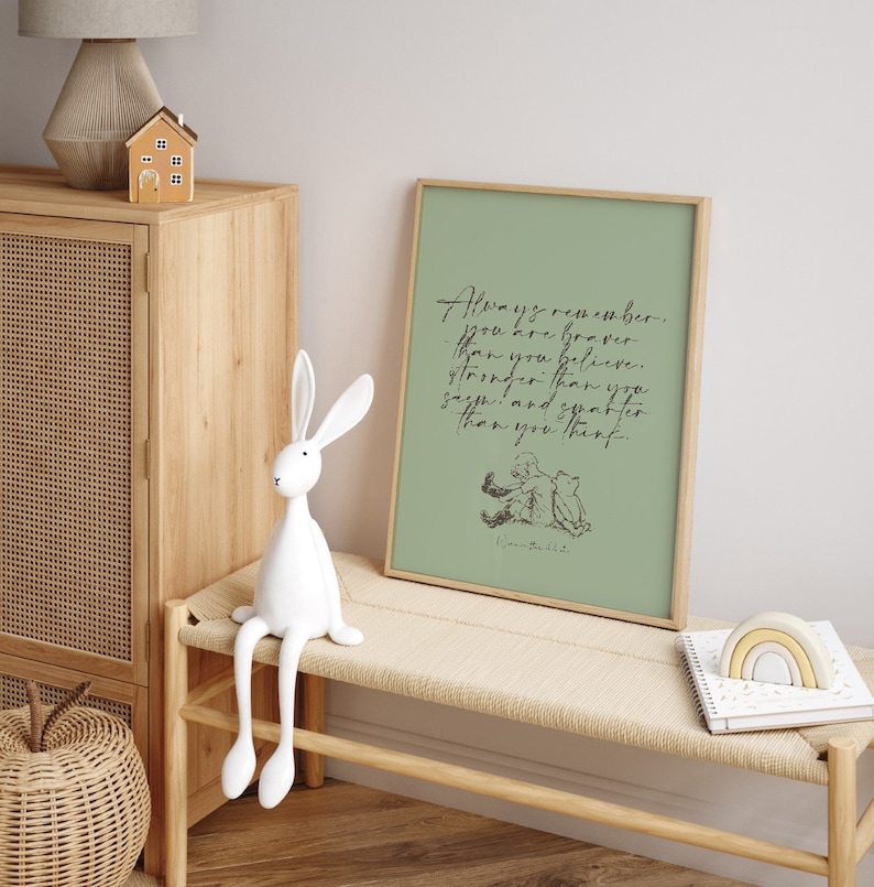 May include: A framed print with a green background and a quote from Winnie the Pooh that reads "Always remember you are braver than you believe, stronger than you seem, and smarter than you think." The quote is in black script font. The print is sitting on a wooden bench with a white rabbit figurine.