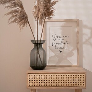 May include: A light wood side table with a woven rattan drawer. A framed print with the text "You are my favourite human" sits on the table. A vase with dried pampas grass sits on the table.