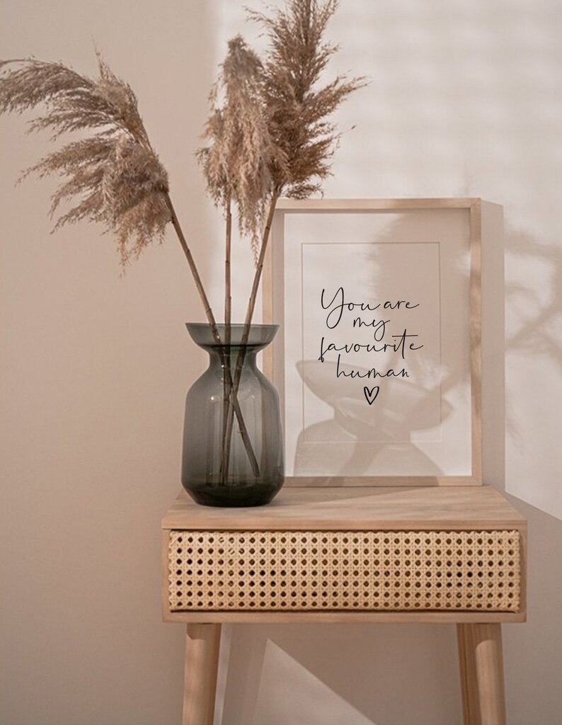 May include: A light wood side table with a woven rattan drawer. A framed print with the text "You are my favourite human" sits on the table. A vase with dried pampas grass sits on the table.
