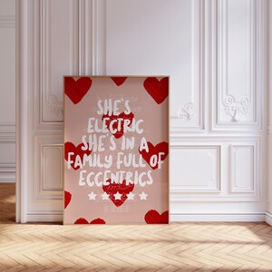 May include: A framed art print with a light pink background, featuring red hearts and white text that reads "She's Electric She's in a Family Full of Eccentrics." The print is in a wooden frame and is displayed against a white wall.