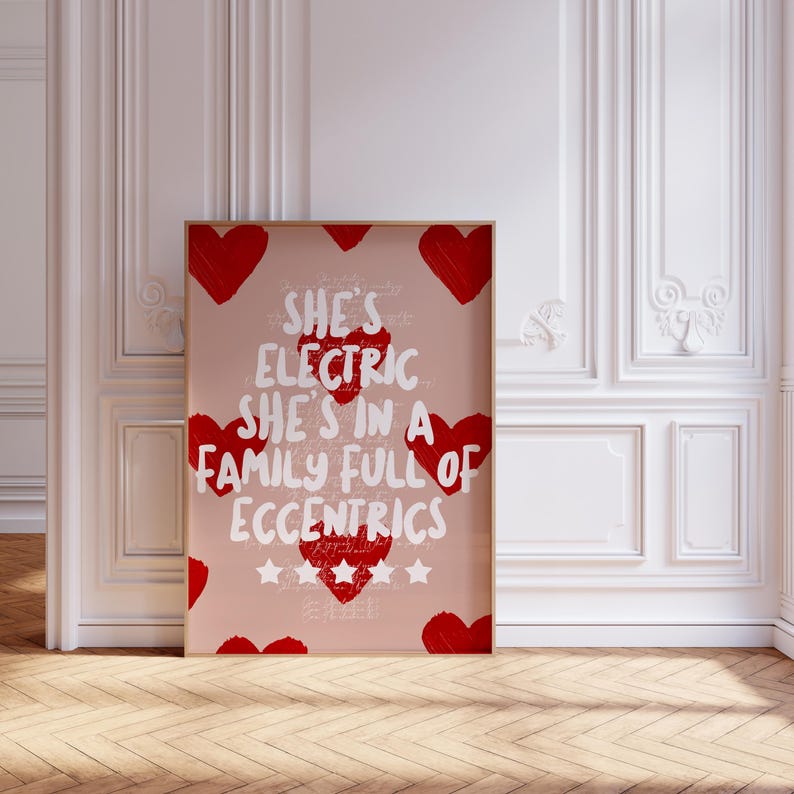 May include: A framed art print with a light pink background, featuring red hearts and white text that reads "She's Electric She's in a Family Full of Eccentrics." The print is in a wooden frame and is displayed against a white wall.