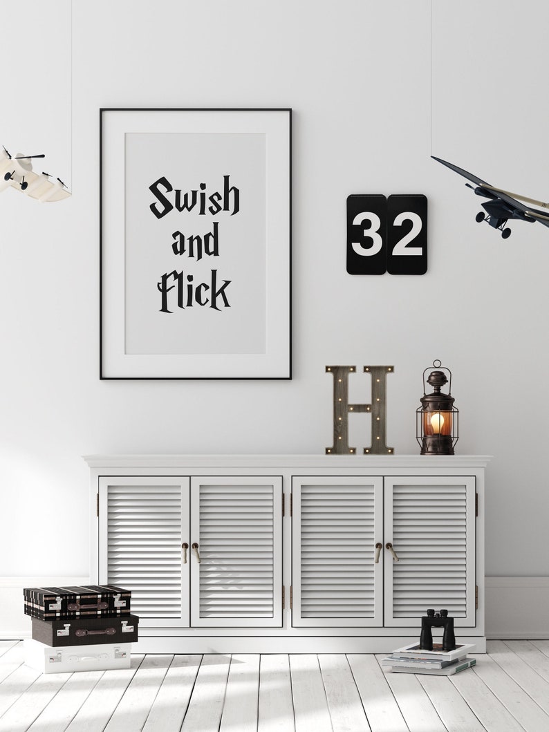 May include: A white cabinet with three doors and a black framed print with the text "Swish and Flick" hanging above it. There is a black metal number "32" hanging on the wall to the right of the print. A black and white letter "H" with lights is on the cabinet. A small black and brown suitcase is on the floor in front of the cabinet. A small toy airplane is hanging from the ceiling above the cabinet.