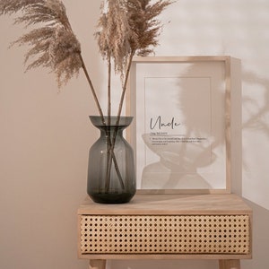 May include: A framed print with the word "Uncle" and a definition of the word. The print is sitting on a light wood table with a woven rattan drawer. A vase with dried pampas grass is on the table.