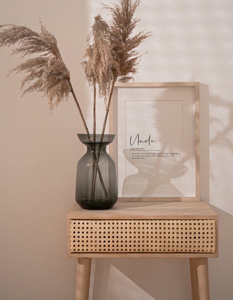 May include: A framed print with the word "Uncle" and a definition of the word. The print is sitting on a light wood table with a woven rattan drawer. A vase with dried pampas grass is on the table.