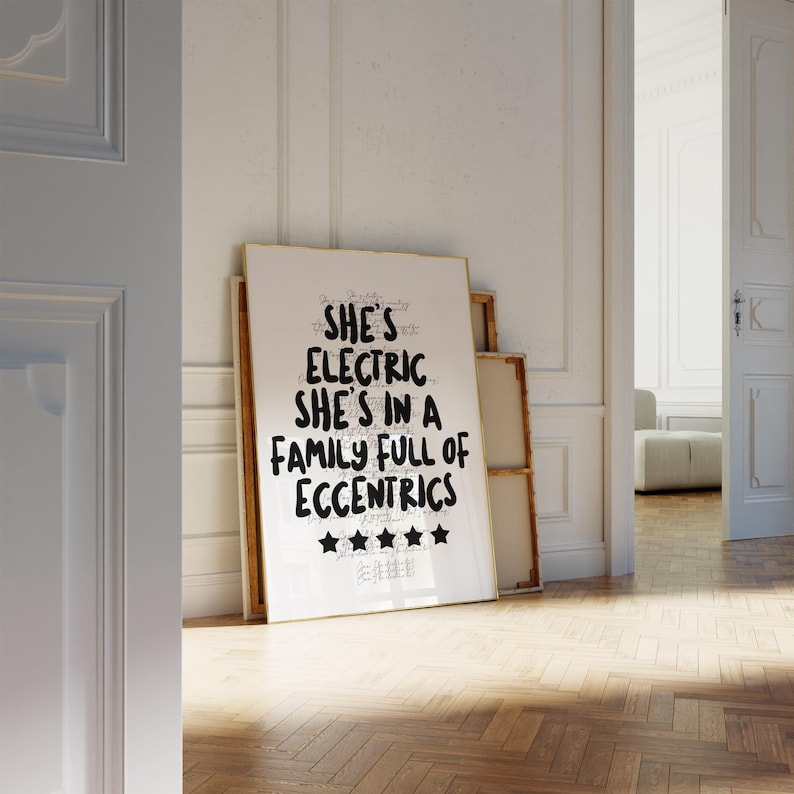 May include: A framed art print with the text "SHE'S ELECTRIC SHE'S IN A FAMILY FULL OF ECCENTRICS" in black lettering. The print is propped against a wall in a room with hardwood floors and white walls.