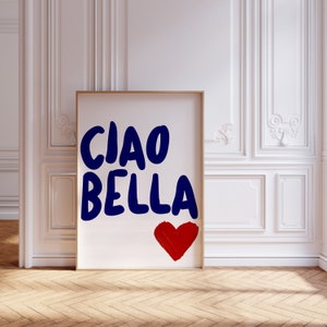 May include: A framed art print with the words "CIAO BELLA" in dark blue, accompanied by a red heart. The artwork is displayed against a white wall with decorative molding and a wooden floor, creating a stylish interior design aesthetic.
