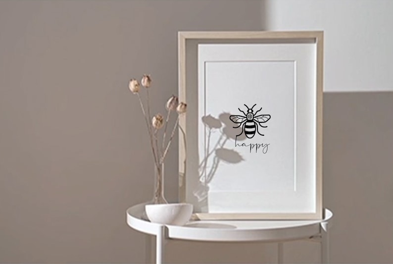 May include: A black and white print of a bee with the word "happy" underneath. The print is framed in a light wood frame and is sitting on a white table.