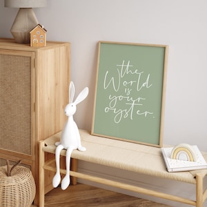 May include: A framed print with a sage green background and white text that reads "The world is your oyster." The print is hanging on a wall above a wooden bench with a white rabbit figurine sitting on it.