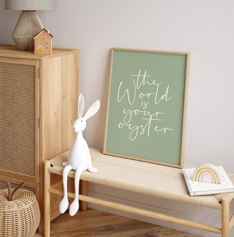 May include: A framed print with a sage green background and white text that reads "The world is your oyster." The print is hanging on a wall above a wooden bench with a white rabbit figurine sitting on it.