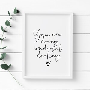 May include: A framed print with the words "You are doing wonderful darling" in black script. A small heart is below the text. The frame is white, and a sprig of green leaves is to the left of the frame.