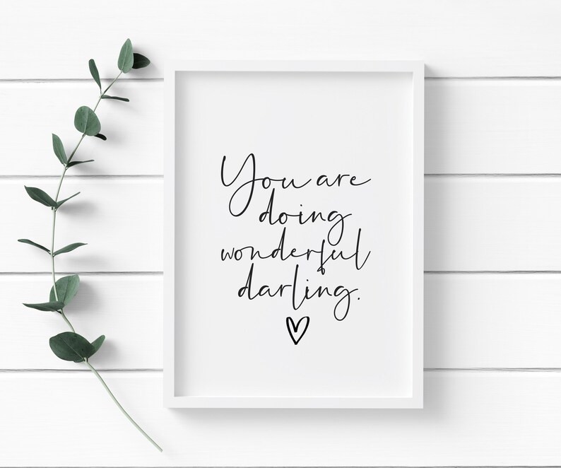 May include: A framed print with the words "You are doing wonderful darling" in black script. A small heart is below the text. The frame is white, and a sprig of green leaves is to the left of the frame.