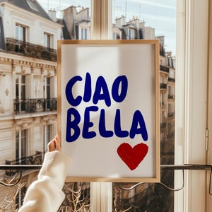 May include: A framed art print with the words "CIAO BELLA" in blue script and a red heart. The artwork is held up in front of a window overlooking a European-style building.