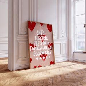 May include: A framed art print with a light pink background and red heart accents. The print features the text "She's Electric She's in a Family Full of Eccentrics" in white. The frame is a light brown color.