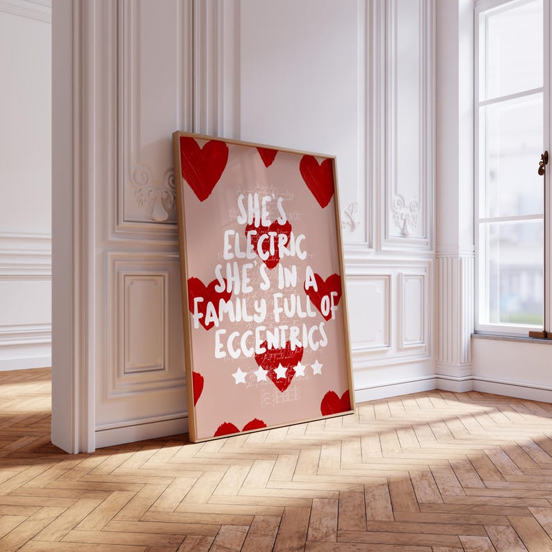 May include: A framed art print with a light pink background and red heart accents. The print features the text "She's Electric She's in a Family Full of Eccentrics" in white. The frame is a light brown color.