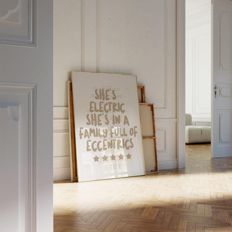 May include: A framed art print with the text "She's Electric She's in a Family Full of Eccentrics" in a light brown font. The print is leaning against a wall in a room with a wooden floor.