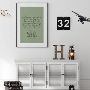May include: A framed print with a green background and a black frame. The print features a quote from Winnie the Pooh: "Always remember you are braver than you believe, stronger than you seem, and smarter than you think." The quote is written in black cursive script. The print also features a black and white illustration of Winnie the Pooh and Piglet sitting together. The print is hanging on a white wall with a black number 32, a wooden letter H, and a small lantern on a white shelf below.