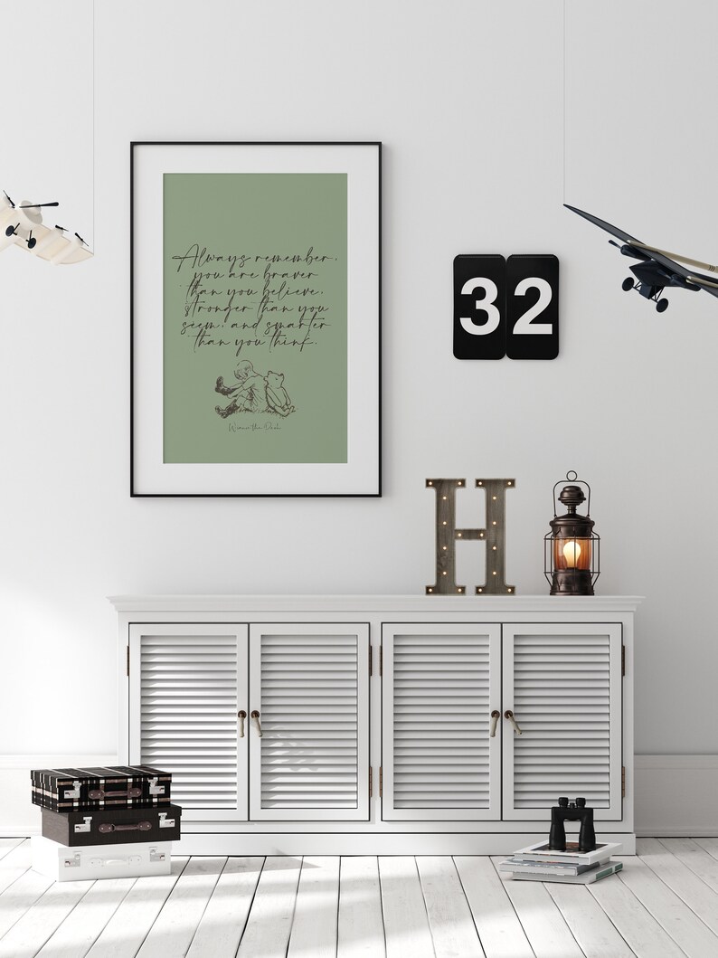 May include: A framed print with a green background and a black frame. The print features a quote from Winnie the Pooh: "Always remember you are braver than you believe, stronger than you seem, and smarter than you think." The quote is written in black cursive script. The print also features a black and white illustration of Winnie the Pooh and Piglet sitting together. The print is hanging on a white wall with a black number 32, a wooden letter H, and a small lantern on a white shelf below.