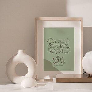 May include: A framed print with a green background and a quote from Winnie the Pooh that reads "Always remember you are braver than you believe, stronger than you seem, and smarter than you think." The print is sitting on a white table with a white vase and a white lamp.