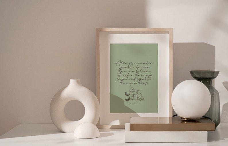 May include: A framed print with a green background and a quote from Winnie the Pooh that reads "Always remember you are braver than you believe, stronger than you seem, and smarter than you think." The print is sitting on a white table with a white vase and a white lamp.