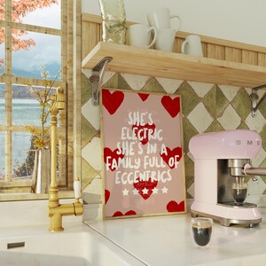 May include: A pink and silver espresso machine with a glass of coffee sits on a white countertop next to a framed print. The print has red hearts and the text "She's Electric She's in a Family Full of Eccentrics."