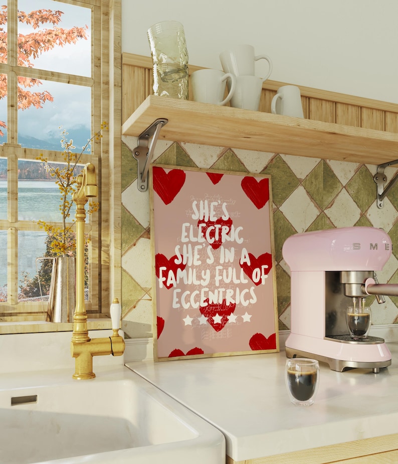 May include: A pink and silver espresso machine with a glass of coffee sits on a white countertop next to a framed print. The print has red hearts and the text "She's Electric She's in a Family Full of Eccentrics."