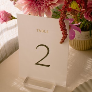 May include: A table number card with the word "TABLE" and the number "2" printed in gold and black. The card is displayed in a clear acrylic holder. A floral arrangement with pink and red flowers is in the background.