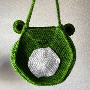 May include: Green crocheted frog-shaped tote bag with a white crocheted circle on the front.