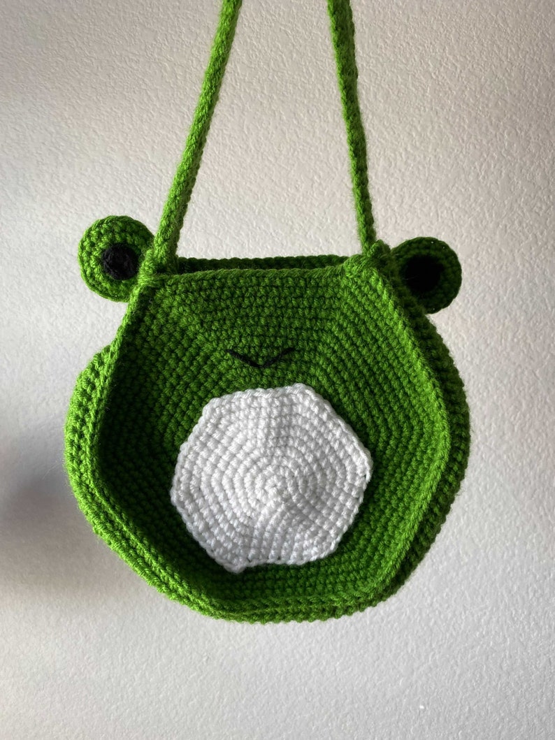 May include: Green crocheted frog-shaped tote bag with a white crocheted circle on the front.