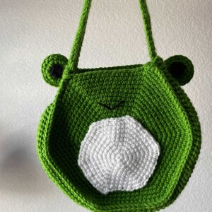 May include: Green crocheted frog-shaped bag with white crocheted details. The bag has two straps and a round shape.