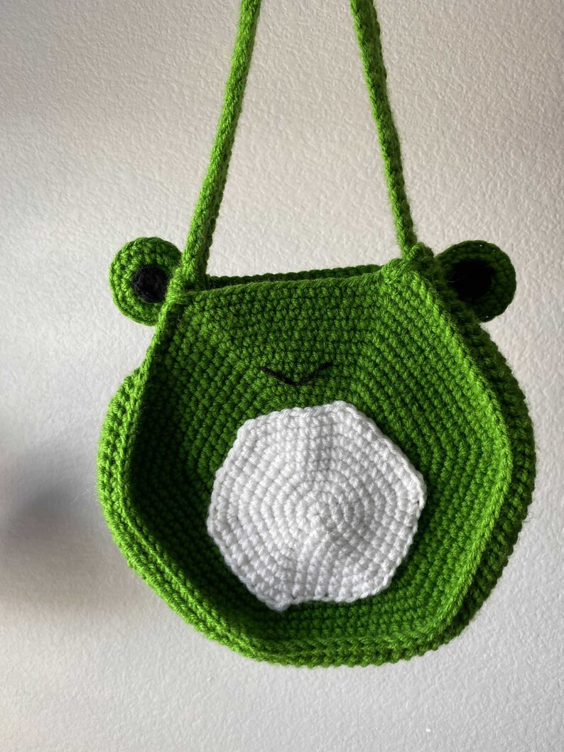 May include: Green crocheted frog-shaped bag with white crocheted details. The bag has two straps and a round shape.