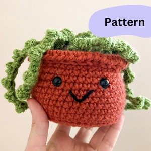 May include: A crocheted plant pot with a smiling face and green crocheted leaves. The pot is brown and the leaves are green.