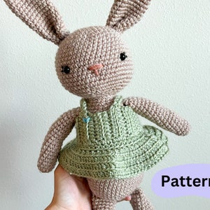 May include: A crocheted bunny wearing a green dress. The bunny is light pink and has black eyes. The dress has a simple design and is a light green color.