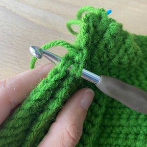 May include: Close-up of a person crocheting with green yarn and a silver crochet hook. The yarn is wrapped around the hook and the person is holding the hook in their right hand.