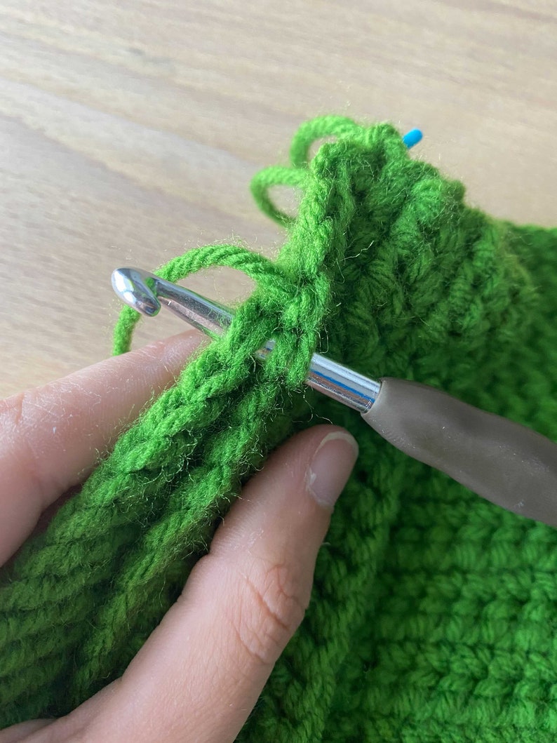 May include: Close-up of a person crocheting with green yarn and a silver crochet hook. The yarn is wrapped around the hook and the person is holding the hook in their right hand.