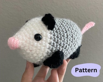 May include: A crocheted plush toy shaped like a possum. The toy is grey, white, and black with a pink nose. The toy is being held in a hand. The text "Pattern" is visible in the image.