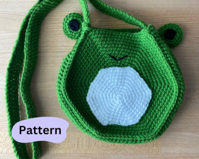 May include: Green crocheted frog-shaped bag with a white crocheted circle in the center. The bag has a long green strap.