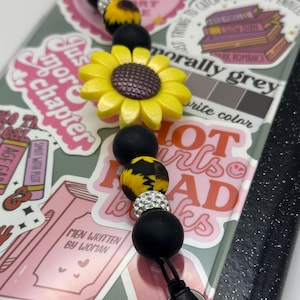 May include: A black and yellow phone charm with a sunflower charm, black beads, and rhinestone accents. The charm is attached to a black phone case with stickers that say "Just More Chapter", "Men Written By Woman", "Hot Girls Read Books", and "Morally Grey".