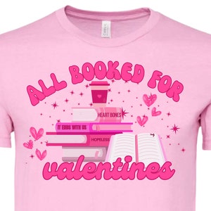 Puede incluir: Camiseta rosa con el texto "All Booked for Valentines" en rosa. El diseño presenta una pila de libros con títulos como "Heart Bones", "It Ends With Us" y "Hopeless", una taza de café y corazones.