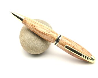 May include: A wooden pen with gold-colored accents rests on a small, smooth, gray stone. The pen has a light brown, wood-grain barrel and a gold-colored clip and tip. The pen is angled slightly, with the tip pointing upwards.