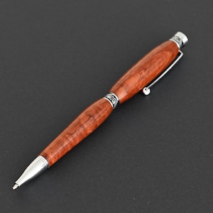 Könnte beinhalten: Ein Kugelschreiber mit einem polierten, rotbraunen Holzgehäuse und sichtbarer Maserung. Der Stift verfügt über silberfarbene Metallkomponenten, darunter eine Spitze, ein dekorativer Mittelring mit Gravur und ein Taschenclip.