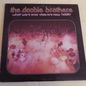 Puede incluir: Una portada de &aacute;lbum de vinilo vintage con una foto en blanco y negro de los Doobie Brothers tocando en el escenario frente a una multitud. El t&iacute;tulo del &aacute;lbum es "What Were Once Vices Are Now Habits".