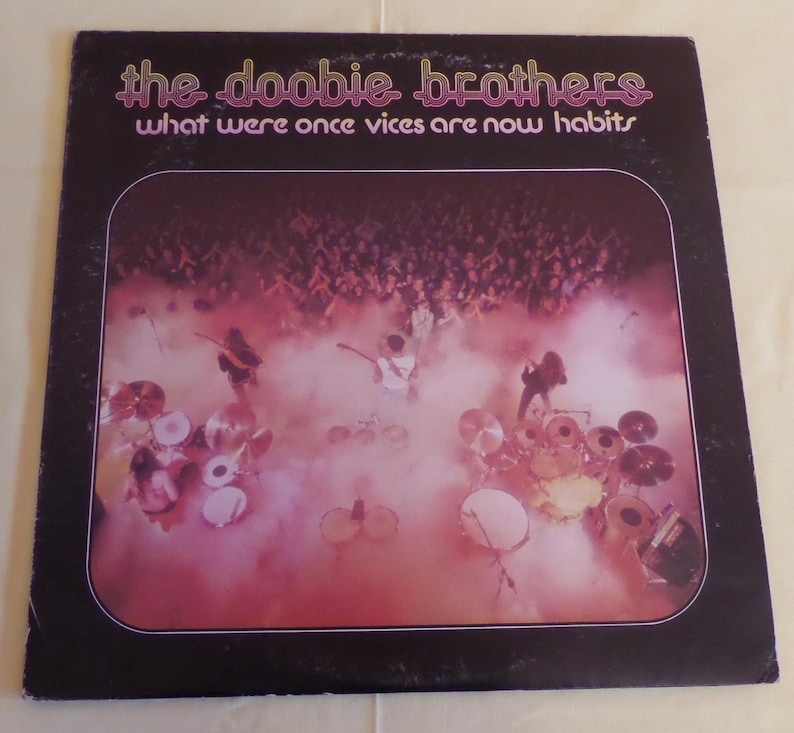 Puede incluir: Una portada de &aacute;lbum de vinilo vintage con una foto en blanco y negro de los Doobie Brothers tocando en el escenario frente a una multitud. El t&iacute;tulo del &aacute;lbum es "What Were Once Vices Are Now Habits".