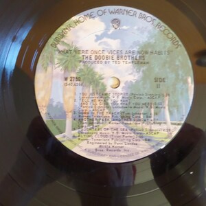 Puede incluir: Un disco de vinilo negro con una etiqueta blanca. La etiqueta tiene un dise&ntilde;o de palmera y texto que dice "Burbank Home of Warner Bros. Records", "What were once vices are now habits", "The Doobie Brothers", "Produced by Ted Templeman", "W 2750", "Side II", y una lista de t&iacute;tulos de canciones.