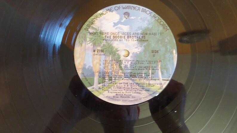 Puede incluir: Un disco de vinilo negro con una etiqueta blanca. La etiqueta tiene un dise&ntilde;o de palmera y texto que dice "Burbank Home of Warner Bros. Records", "What were once vices are now habits", "The Doobie Brothers", "Produced by Ted Templeman", "W 2750", "Side II", y una lista de t&iacute;tulos de canciones.
