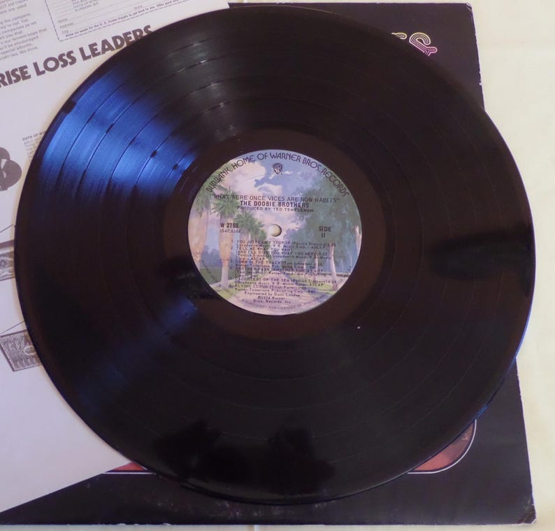 Puede incluir: Disco de vinilo negro con una etiqueta blanca. La etiqueta tiene el texto "What Were Once Vices Are Now Habits" y "The Doobie Brothers" impreso en ella. El disco es de Warner Bros. Records.