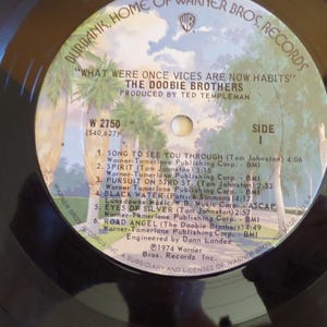 Puede incluir: Un disco de vinilo negro con una etiqueta blanca. La etiqueta tiene el texto "What Were Once Vices Are Now Habits" y "The Doobie Brothers". El disco fue lanzado en 1974 por Warner Bros. Records.