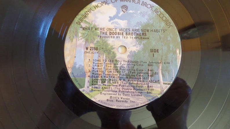 Puede incluir: Un disco de vinilo negro con una etiqueta blanca. La etiqueta tiene el texto "What Were Once Vices Are Now Habits" y "The Doobie Brothers". El disco fue lanzado en 1974 por Warner Bros. Records.