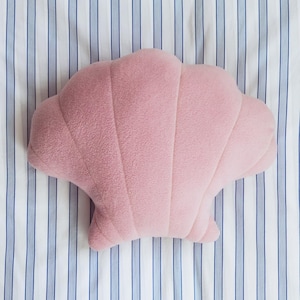 Può includere: Un cuscino rosa a forma di conchiglia con una superficie strutturata. Il cuscino è appoggiato su uno sfondo a righe bianche e blu. Il design presenta linee verticali e curve che imitano le creste di una conchiglia.