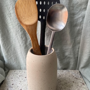 Pode incluir: Um porta-talheres de cerâmica bege com manchas com três utensílios de cozinha dentro: uma colher de madeira, uma colher ranhurada preta e uma colher de servir prateada.