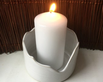 May include: A white ceramic candle holder with a wavy edge, holding a white pillar candle with a lit flame.